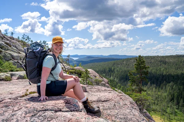Caucasian outdoor active young adult woman sitting on Rock With outdoor hiking clothes and cap smiling and looking at camera with mountain and lake background in Skuleskogen national park, Sweden.