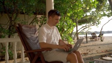 Young businessman on tropical island works at a laptop and looks into the camera. The concept of a pleasant rest and stay compatible with online work