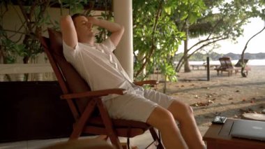 A man rests on a chair on the shores of the Indian Ocean, wearing glasses. The concept of a sleep break while working on a tropical island
