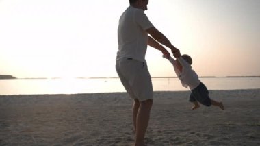 Wide shot of father spins his child by hands on the ocean hotel beach. Funny game of toddler and his caucasian father.