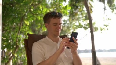 A young man in search of information on his phone on a tropical island close-up. Caucasian businessman finishes working with smartphone puts on glasses and begins to rest