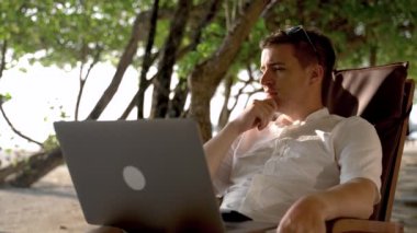 The pensive businessman looks towards the beach and the ocean and continues to work at the laptop. Vacation on Maldives hotel with duty to online working.