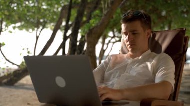 A young businessman on vacation works at a laptop outdoor. The guy looks towards the ocean and reflects on his work.