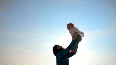 Mother cheerfully throws a small child into the sky in her arms. Happy toddler smiles and laughs flying past the dawn sun