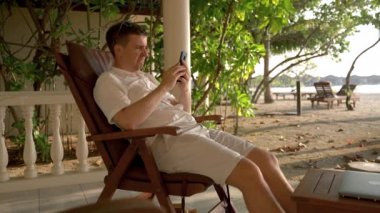 A businessman man sitting on the beach fails. He begins to bang his head against the sunbed, his glasses fall on his eyes, defeats at work online.
