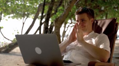 The snide hater came up with an unpleasant comment and writes it on the social network. Beach, sand, palm trees in the background. A young man on vacation watches the content rubbing his hands.