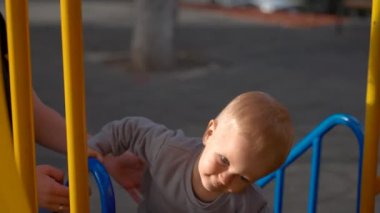 Blond boy climbs colorful metal structure installed on playground in park. Mom helps cute toddler to climb up on blurred background closeup