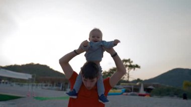 Father entertains little son carrying on shoulders at coastal resort. Man and cute toddler enjoy walking on beach after sunset low angle shot slow motion
