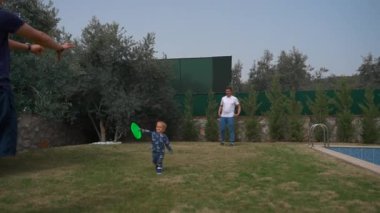 Toddler walks holding frisbee and giving to father. Men and cute little boy play game near swimming pool in yard of country house slow motion