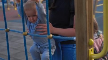 Mother holds little son trying to climb on metal construction installed at playground. Woman entertains toddler in attraction park closeup
