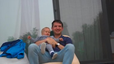 Father talks to little son sitting in comfortable armchair near house with big panoramic windows. Cute blond toddler sits in arms of young man