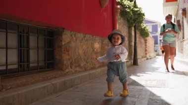Toddler in hat walks with mom on narrow street with old vintage buildings. Cute little boy enjoys travel in touristic town on sunny summer day slow motion