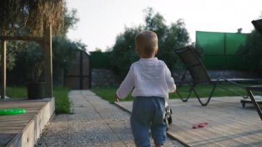 Little boy walks on road towards scooter standing in yard of country house. Blond toddler spends summer vacation in countryside backside view slow motion