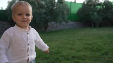Cute little boy in white shirt walks in yard of country house at bright sunlight. Blond toddler enjoys spending time in countryside closeup slow motion