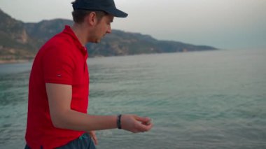 Man throws pebble into sea to jump on water surface at sunset. Tourist stands on beach against mountains enjoying summer vacation closeup slow motion