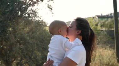 Young mom kisses little son walking in garden with lush greenery on sunny day. Brunette woman and blond toddler enjoy nature at rural site closeup slow motion