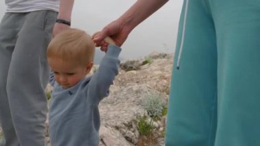 Blond toddler holds hands of parents walking on rocky hill against distant mountains. Family hikes exploring new tourist place closeup slow motion