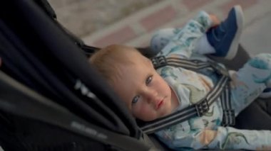 Blond boy rides in stroller fastened with seat belt on blurred background. Toddler looks at camera enjoying walk in city park with parents closeup