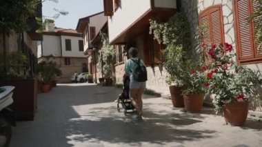 Woman walks with stroller on street past pot-plants standing against vintage buildings. Mother and child on trip in touristic town on sunny day back view