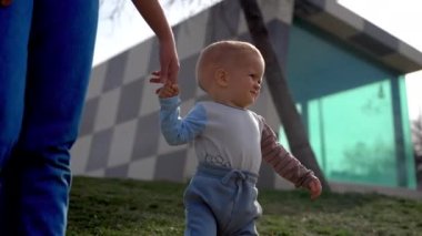 Toddler boy joins hands with father walking on green grass against creative building. Caring parent teaches little son to walk closeup slow motion