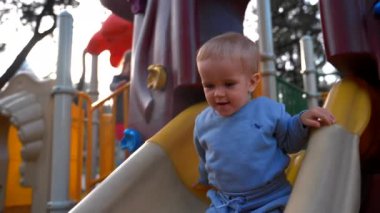 Little kid boy slides down colorful slide installed at playground in urban park on sunny day. Cute toddler enjoys spring activity closeup slow motion