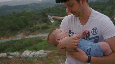 Dad rocks baby girl in arms against mom with bottle of water standing on high hill. Young family with kid explores nature closeup slow motion
