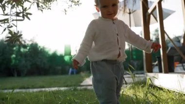 Curious boy walks barefoot on lush grass near tree branches at bright back sunlight. Toddler enjoys sunny summer day in yard closeup slow motion