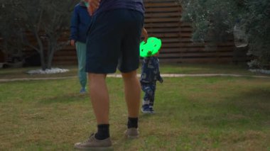 Cute blond boy gives father bright green frisbee . Men and toddler son play fun game in backyard of country house on sunny spring day slow motion