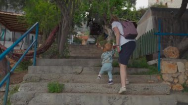 Mother with backpack helps toddler son to get up stairs near lush green trees. Woman and little blond boy explore new town on vacation backside view