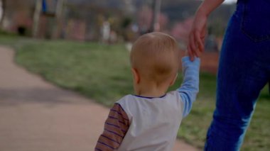 Father holds hand of son walking on wooden road in city park on sunny day backside view. Man and toddler on blurred background closeup slow motion
