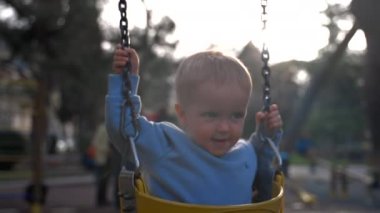 Toddler rides swings holding on to iron chains on blurred background. Blond boy enjoys sunny day at playground in city park closeup slow motion