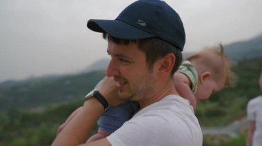 Father puts daughter on shoulder turning to mother standing on high hill against mountains. Man and baby girl on blurred background closeup slow motion