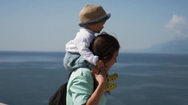 Mother carries toddler son on shoulders walking on waterfront overlooking sea near giant mountains. Woman and kid visit coastal town closeup slow motion