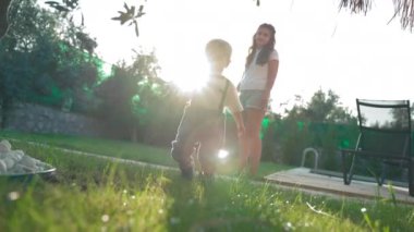 Sweet boy walks on lush grass at sunset light in yard with green trees in summer. Toddler and mother spend weekend in country house slow motion