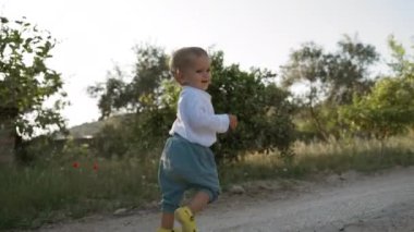 Little child walks on ground road against village garden lush trees casting shadows in summer. Toddler boy explores rural nature in countryside backside view