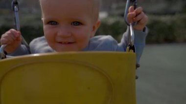 Cheerful boy rides swings on blurred background. Blond toddler enjoys activity at playground for children on sunny day closeup slow motion