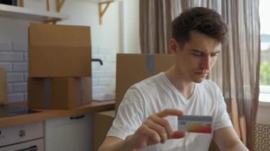 Young man buys domestic appliances on website via laptop typing payment details. Home owner sits at kitchen table against paper boxes close view