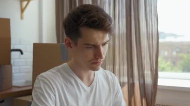 Focused man finishes report on laptop sitting at kitchen table against packed paper boxes. Freelancer takes time for work after relocation closeup