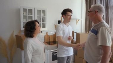 Elderly parents give key to new apartment to married son standing in kitchen. Happy young man shakes hands with dad and hugs mom feeling thankful for gift