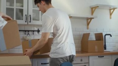 Happy couple moves to new apartment bringing things in paper boxes and putting on countertop. Young man and woman sit at kitchen table smiling closeup