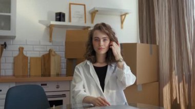 POV of young woman fixing hairstyle with hand looking in camera. Lady sits at kitchen table near closed laptop against paper boxes in new apartment slow motion