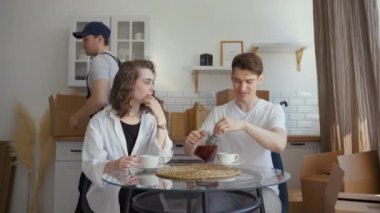 Husband pours hot tea into cups sitting with wife at kitchen table of new apartment. Courier helps young couple with relocation carrying heavy cardboard boxes