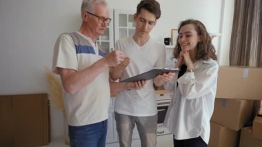 Elderly realtor discusses purchase of apartment with young couple standing in kitchen. Husband signs agreement papers while brunette wife feels excited