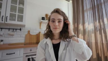 POV of young woman fixing hairstyle with hand looking in camera and smiling. Lady sits at kitchen table at home during online conference closeup