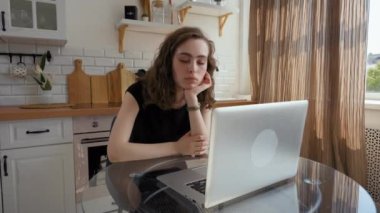 Tired young woman falls asleep while working on laptop in kitchen. Magic hand pops out of screen of electronic device trying to wake up sleepy lady