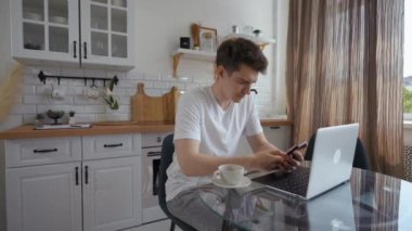 Young man types message on phone sitting near laptop at glass table in modern kitchen. Busy guy distracts on gadget while working online at home