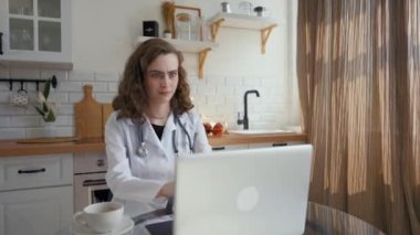 Young lady doctor grimaces looking at unpleasant moments on online video courses on laptop. Woman improves qualification studying at home in kitchen