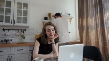 Woman talks on video call with girl friend against man washing dishes in sink. Young lady sits at glass table near modern laptop in kitchen at home