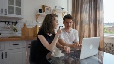 Young engaged couple discusses upcoming wedding with friends on video call via laptop. Happy man and woman sit at at kitchen table looking at screen of device