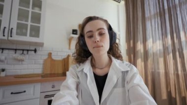 POV of young lady sitting at kitchen table at home looking in camera. Woman in headphones enjoys listening favorite song at break from work closeup